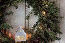 an evergreen holiday wreath with pinecones, lights and a white house with light inside is a cozy and cool idea
