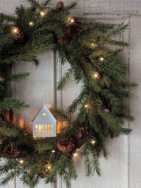 an evergreen holiday wreath with pinecones, lights and a white house with light inside is a cozy and cool idea