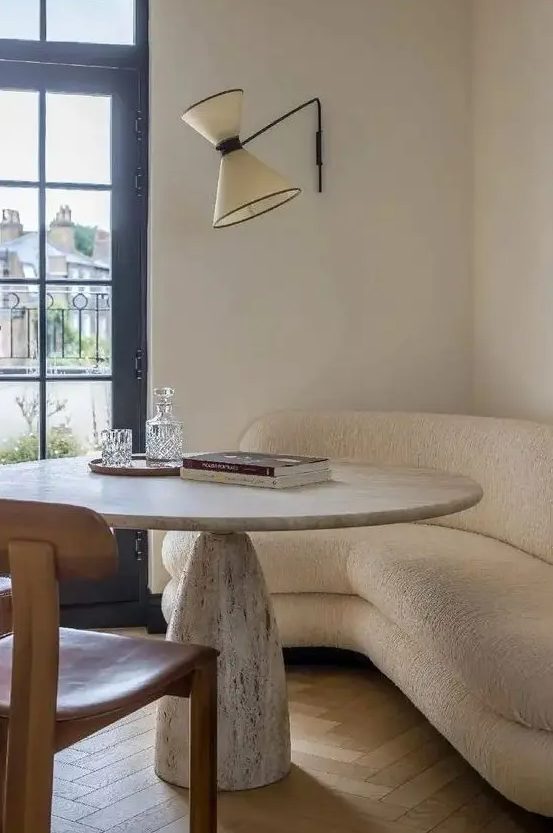 a neutral curved sofa, a stone table, a leather chair and a sconce are a cool combo for a dining nook