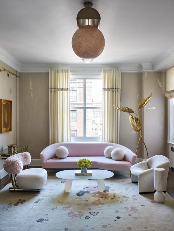 a neutral living room with tan walls, a curved pale pink sofa and curved chairs, a spotted rug and whimsical decor