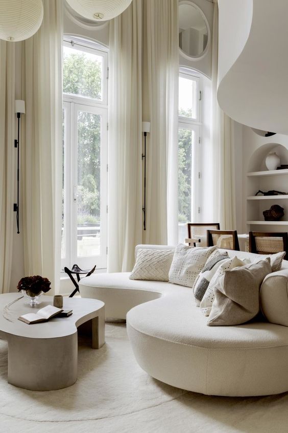 a neutral living room with a creamy curved sofa and pillows, a low coffee table and some lamps and curtains