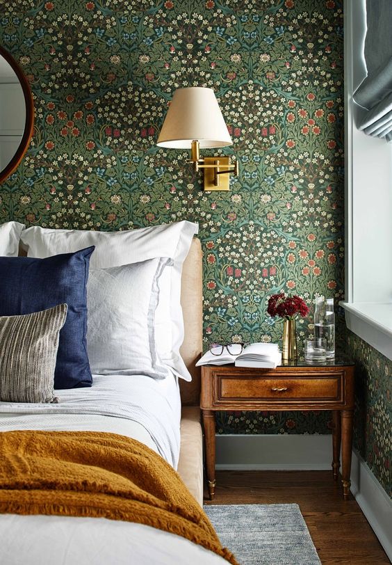a classic English bedroom with vintage floral wallpaper, an upholstered bed with neutral bedding, a stained nightstand and a sconce