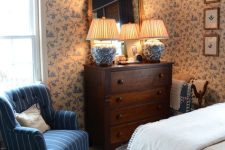 19 a vintage English bedroom with a bed and neutral bedding, a dark-stained dresser, a blue chair, lovely blue wallpaper