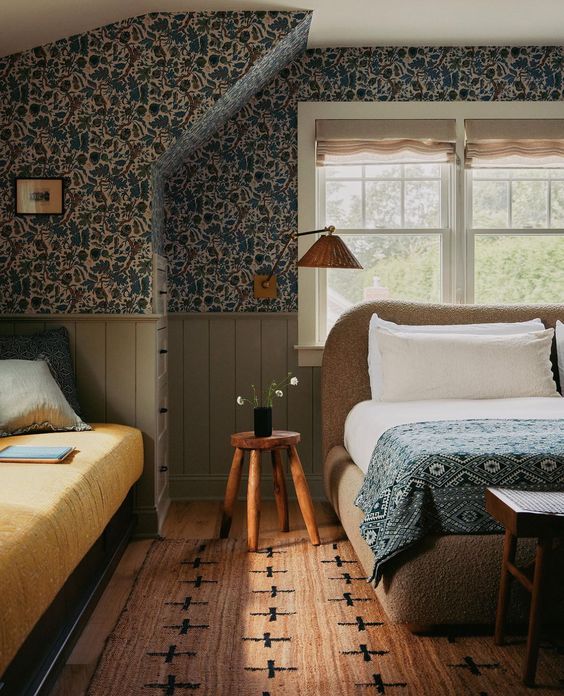 a vintage attic bedroom with dark floral wallpaper, a boucle upholstered bed with printed bedding, a rug and a sofa at the window