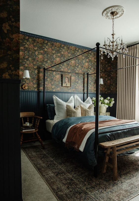a moody vintage bedroom with dark floral wallpaper, a metal canopy bed, navy paneling and bedding, a crystal chandelier