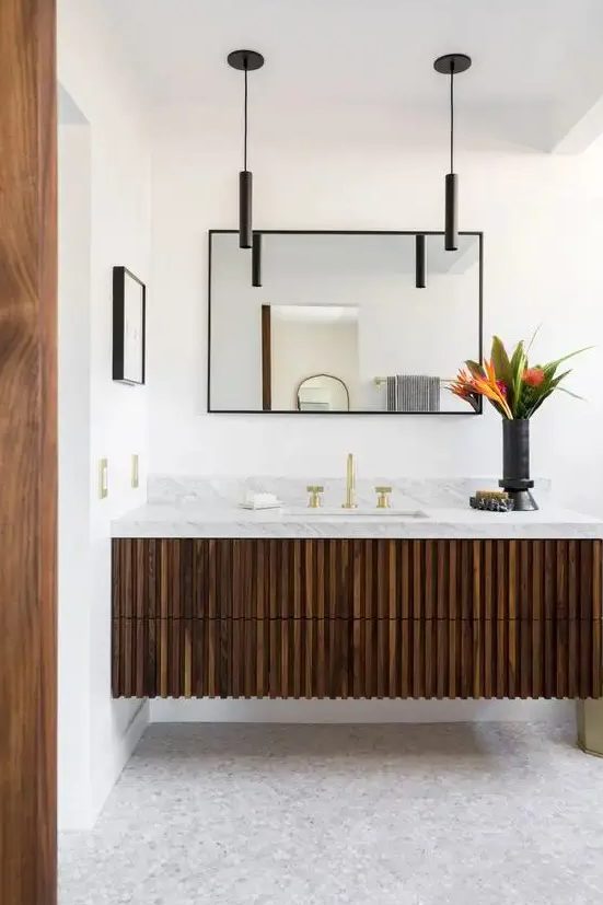 a dark-stained fluted vanity with a stone countertop, a mirror in a black frame, black pendant lamps and bold blooms