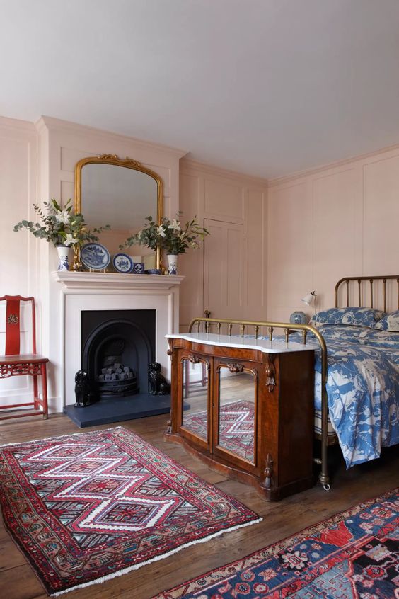 a blush bedroom with a fireplace, a metal bed with printed bedding, a mirror cabinet, colorful printed rugs and blooms