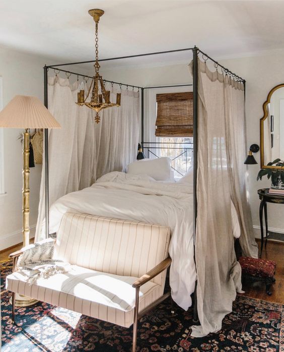 a welcoming vintage bedroom with a cnaopy bed and neutral bedding, a striped loveseat, a floor lamp and a gilded chandelier