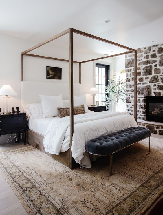 a modern farmhouse bedroom with a stone fireplace, a wood canopy bed, a black tufted bench, black nightstands
