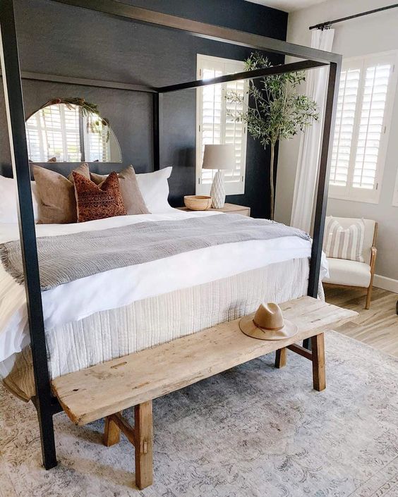 a modern farmhouse bedroom with a black accent wall, a black canopy bed, a stained bench, a potted tree and a creamy chair