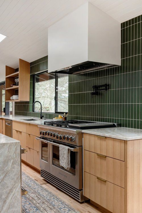 a stained mid-century modern kitchen with a green stacked tile backsplash, a large hood and a stone kitchen island