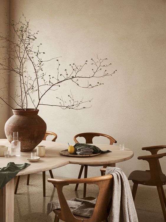 a warm neutral dining room with a catchy table, stained chairs, a pot with branches and some textiles