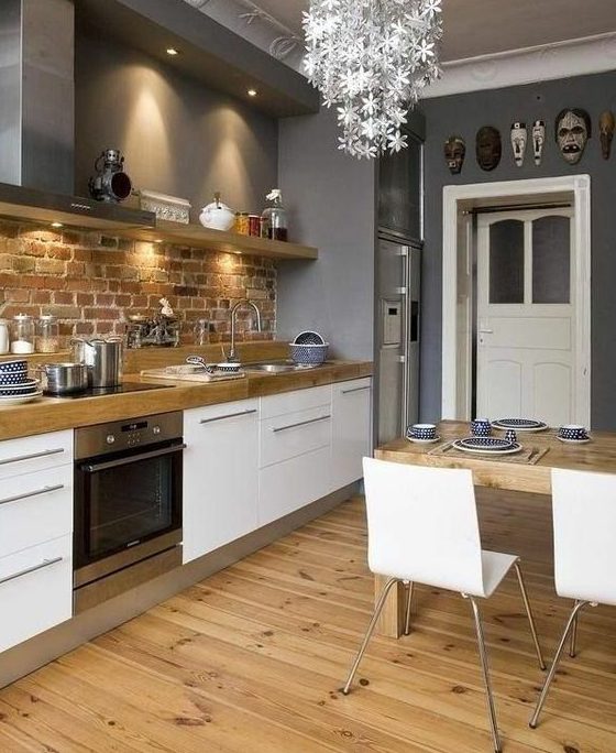 a stylish Scandinavian kitchen with white cabinets, wooden countertops, a brick backsplash, a floral chandelier, white chairs and a wooden table