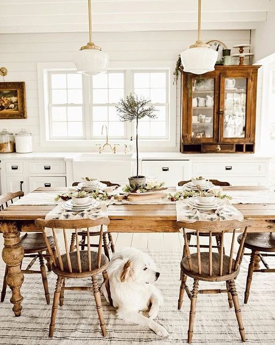 a white farmhouse kitchen with vintage wooden cabinets, a vintage wooden table, chairs and pendant lamps plus artworks