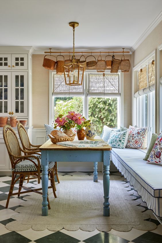 a white cottage kitchen and a corner dining zone with a bench, a blue table and rattan chairs, baskets hanging over the window