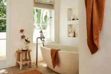 a beautiful earthy bathroom with white plaster walls, a parquet flooring, a stone bathtub, niches with decor and rust textiles
