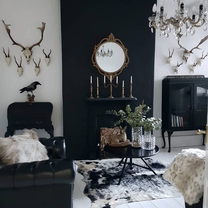 a black and white modern Goth living room with a black fireplace, a black sofa, neutral pillows, an armoire, a table with greenery and skulls