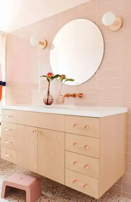 a blush bathroom with skinny tiles, a stained floating vanity, clear glass handles, a pink stool and a terrazzo floor