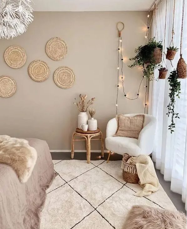 a boho bedroom with taupe walls, a bed, a creamy chair, a rattan stool, hanging potted greenery, decorative woven plates and layered rugs