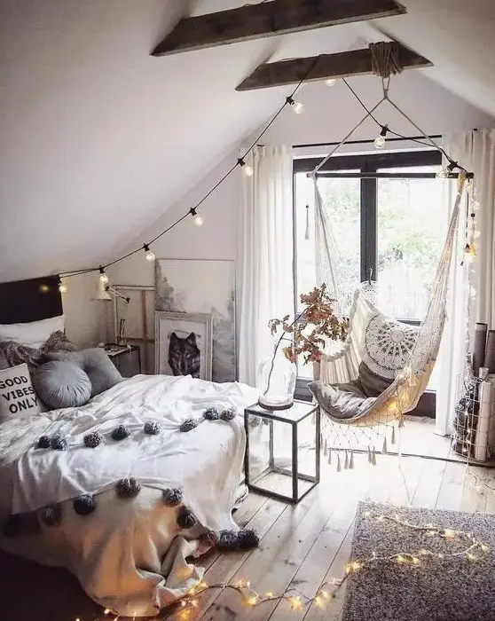 a boho teen attic bedroom with a bed with neutral bedding, a woven pendant chair, lights, wooden beams and artwork