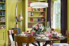 a bold chartreuse dining room with built-in shelves, a stained table and chairs, a pendant lamp and some books
