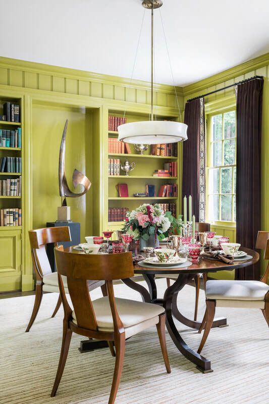 A bold chartreuse dining room with built in shelves, a stained table and chairs, a pendant lamp and some books