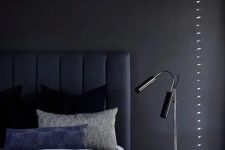 a chic and peaceful moody bedroom with a soot accent wall, a navy upholstered bed and blue bedding, a nightstand, black table lamps