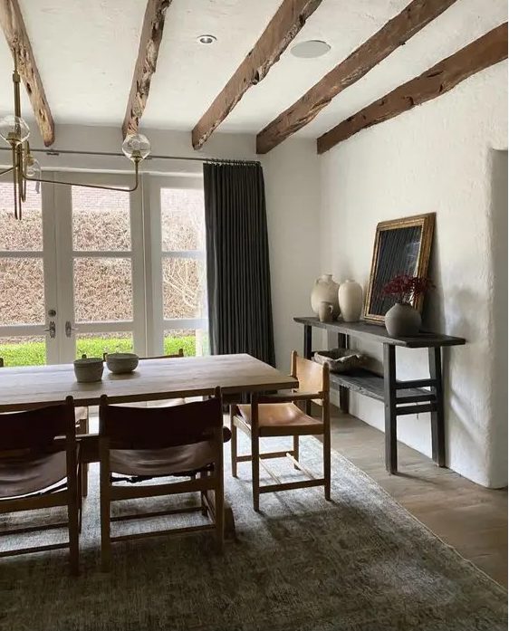 a chic earthy dining room with a glazed wall, a large stained table and leather chairs, a printed rug, a black console table and wooden beams