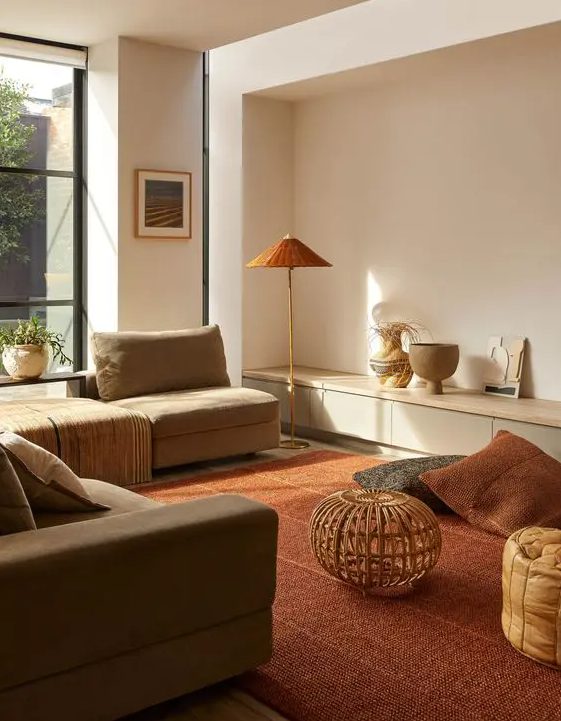 a contemporary warm earthy living room with a grey sofa and neutral seats, a deep red carpet and pillows, a floor lamp and an amber pouf