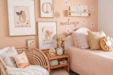 a cozy kid’s room with a peachy accent wall, a bed with pastel bedding, a rattan chair and table and some decor