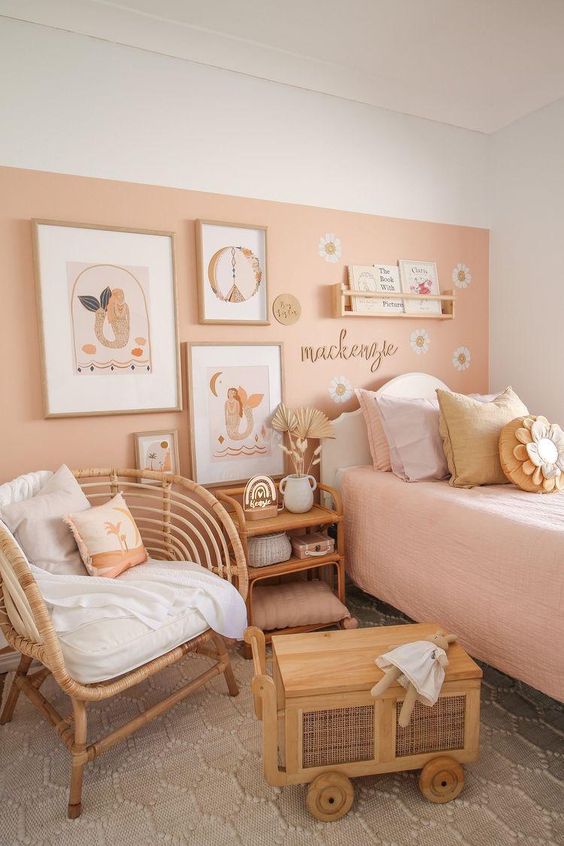 a cozy kid's room with a peachy accent wall, a bed with pastel bedding, a rattan chair and table and some decor