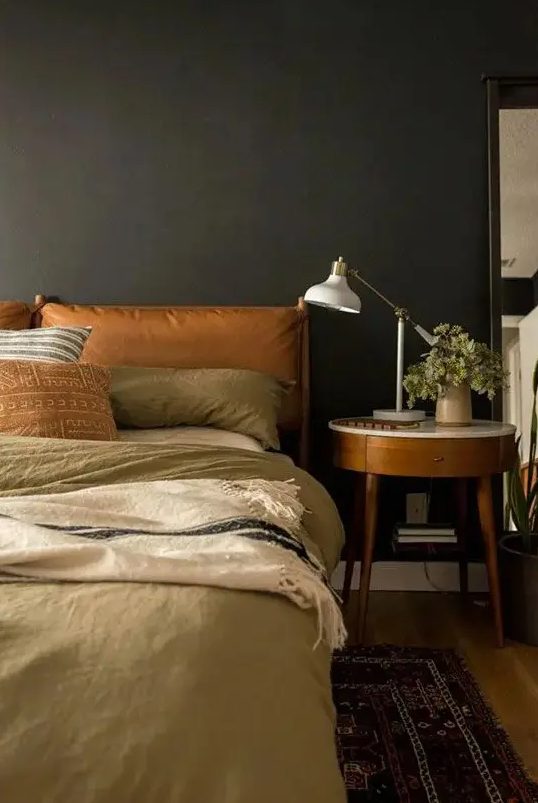 a cozy moody bedroom with soot walls, a bed with amber leather upholstery, neutral bedding, a chic nightstand and a printed rug