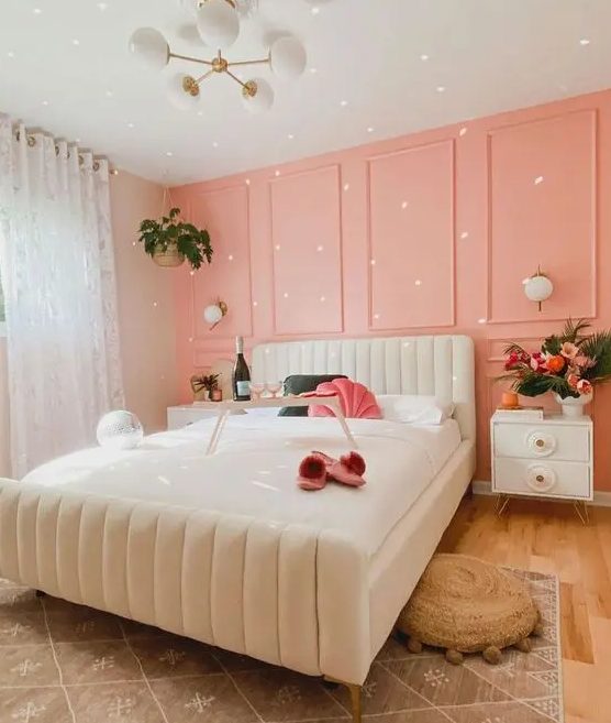 a dreamy bedroom with a peachy pink paneled accent wall, a creamy bed and nightstands, a chandelier and layered rugs