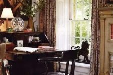 a fab vintage home office with vintage artwork, a dark-stained bureau desk and a black chair, printed curtains and a rug plus some blooms