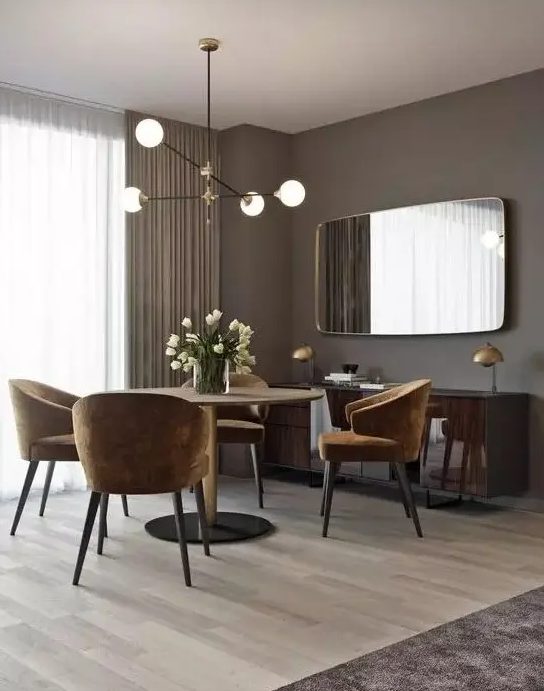 A glam and chic taupe dining room with a round table, rust colored chairs, a glossy floating credenza and a large mirror