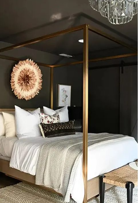 a glam moody bedroom with soot walls and a ceiling, a gold canopy bed with neutral bedding, a woven bench and a crystal chandelier