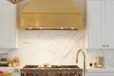 a glam white kitchen with shaker cabinets, a grey kitchen island, a white stone backsplash and countertops and a gold hood
