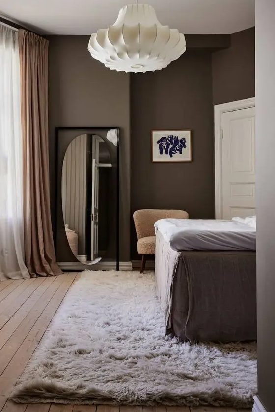 a laconic bedroom with brown walls, a tall bed, a neutral rug, a floor mirror, mauve curtains and a lamp