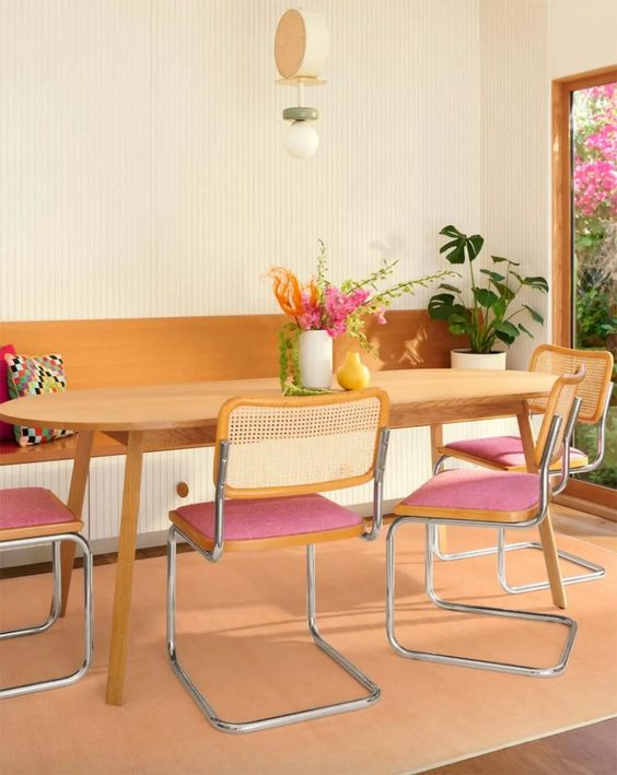 A lively dining room with a buil in bench, a table, pink chairs with cane backs, a Peach Fuzz rug and some plants