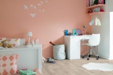 a lively kids’ room with a Peach Fuzz accent wall, a white bed and printed bedding, a white desk and a chair and peachy shelves