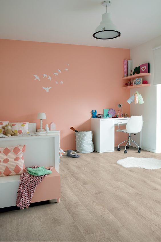 a lively kids' room with a Peach Fuzz accent wall, a white bed and printed bedding, a white desk and a chair and peachy shelves