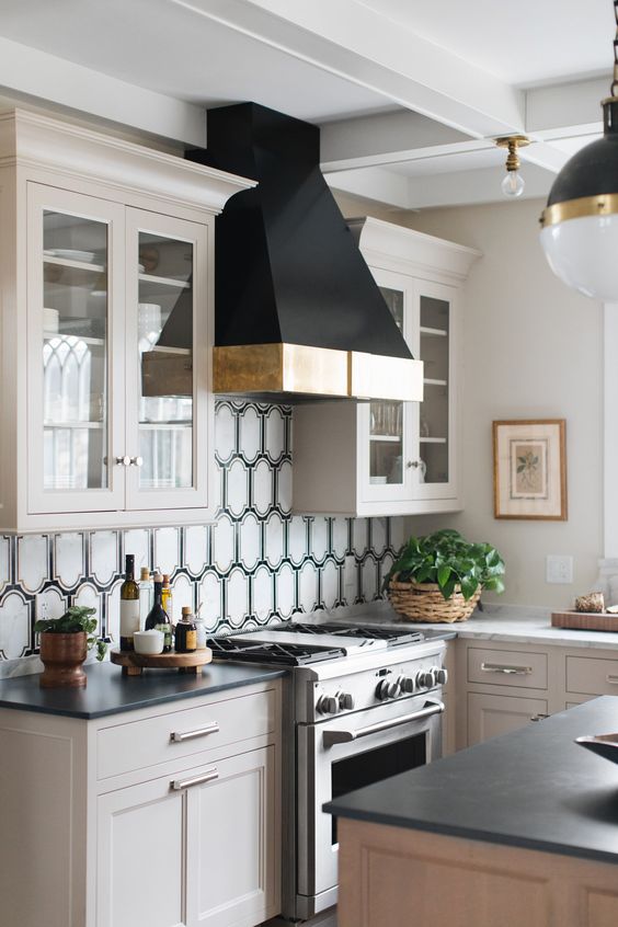 a lovely farmhouse kitchen with light-stained cabinets, grey stone countertops, a whimsical backsplash and a large black hood