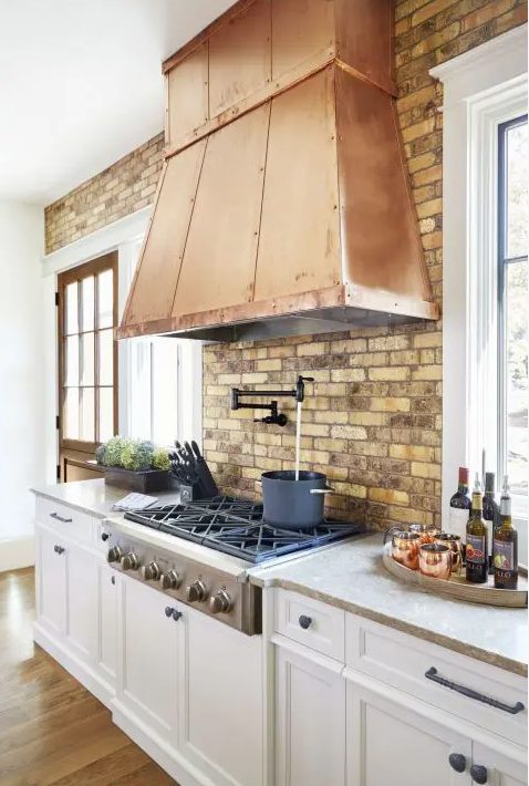 a lovely white farmhouse kitchen with shaker cabinets, a brick backsplash and a large copper hood that adds a warm touch