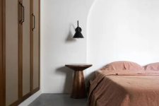 a minimal earthy bedroom with a built-in wardrobe, a bed with rust bedding, a metal nightstand and a black sconce