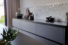 a minimalist sleek grey kitchen with only lower cabinets, a white fishscale tile backsplash and a built-in hood over the space