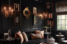 a modern Goth living room with black walls and a ceiling, a black sofa, gold pillows, a neutral ottoman, a gallery wall and a chandelier