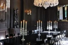 a modern Gothic dining room with black panel walls, a glossy table, upholstered chairs, a large white chandelier and artworks