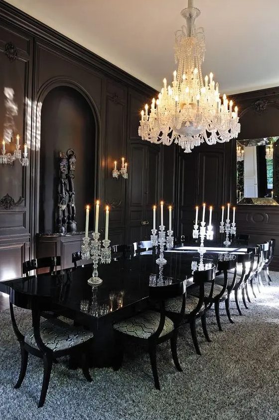 a modern Gothic dining room with black panel walls, a glossy table, upholstered chairs, a large white chandelier and artworks
