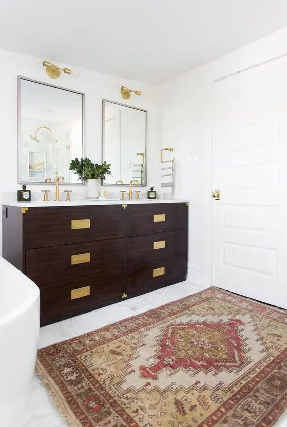 a modern farmhouse bathroom with a double dark-stained vanity with gold fixtures and gold handles, mirrors and gold sconces over them