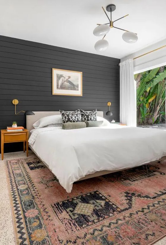 a modern farmhouse bedroom with a black shiplap accent wall, a neutral bed, a printed rug, light-stained nightstands and a cool chandelier